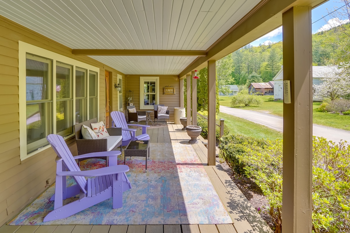 A spacious front porch is displayed, featuring colorful lavender Adirondack chairs and a patterned rug. Overhead, a ceiling is white with a wooden facade. Surrounding lush greenery enhances the outdoor relaxation space, while a pathway leads to the road beyond.