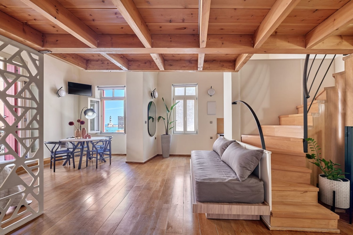 The main living area features a minimalist design with wooden flooring and exposed beams. Ample natural light enters through large windows, highlighting a grey sofa and a dining table for four. The staircase leads to the upper level, enhancing the spacious feel of the room.
