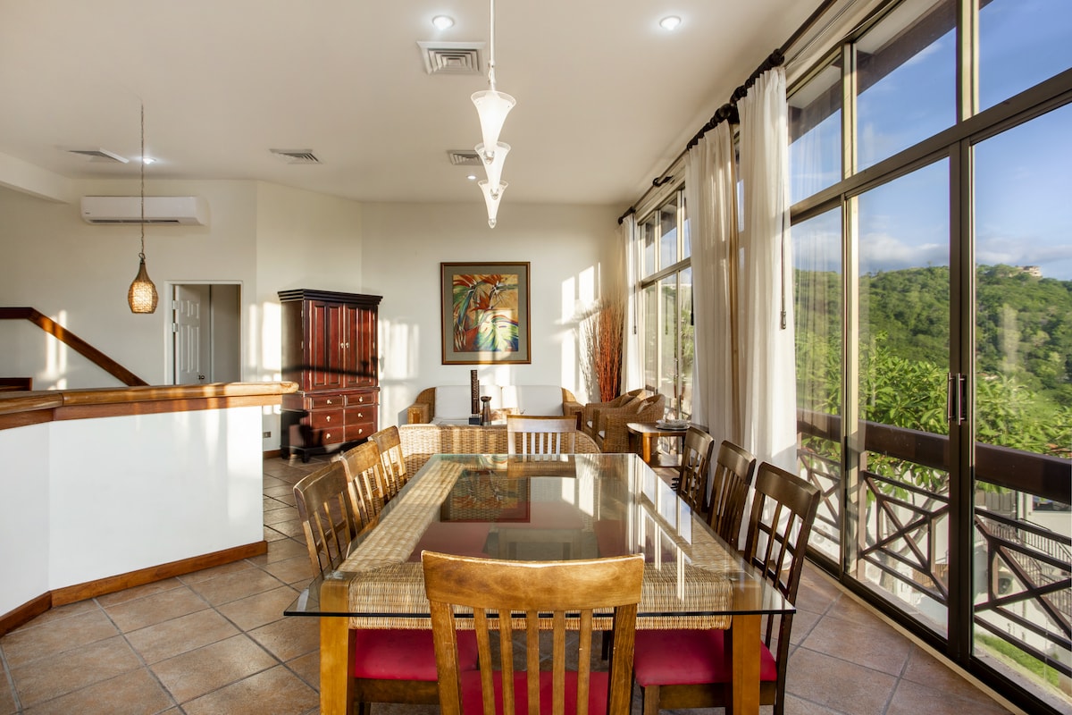 A spacious dining area is highlighted by a large glass table surrounded by wooden chairs. Natural light flows through expansive windows, offering views of the greenery outside. A vibrant piece of art adorns the wall, complemented by soft lighting fixtures hanging from the ceiling.