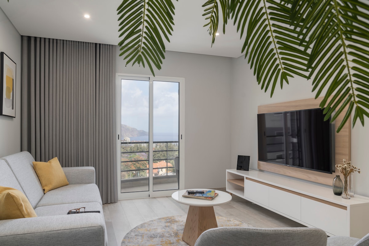The living area features a light grey sofa complemented by a mustard yellow cushion. A round coffee table occupies the center, with a modern TV mounted on a light wood storage unit. Sliding glass doors provide access to the balcony and a view of the surrounding landscape.