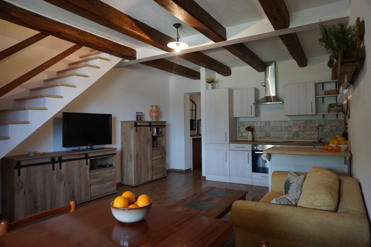 The living area is enhanced by natural wood beams and features a staircase leading to the upper level. A TV is mounted on a wall unit, and a fully-equipped kitchen is visible. A dining table with a bowl of oranges and a cozy sofa complete the space.