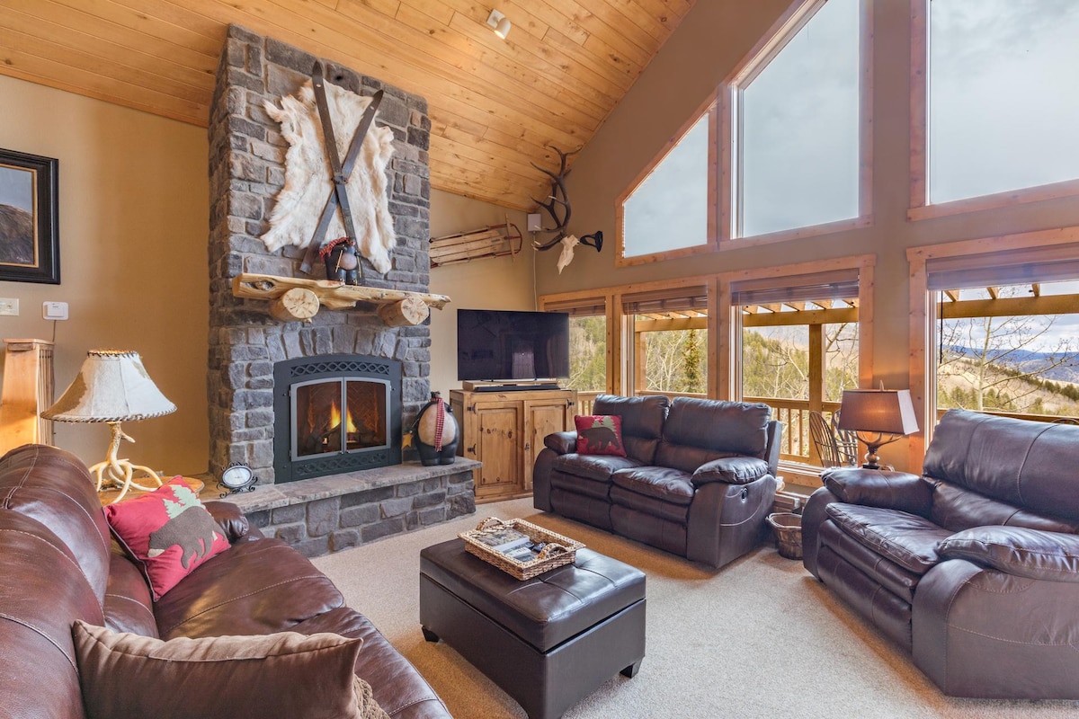 A spacious living area features large windows that offer views of the surrounding landscape. A stone fireplace is centered against a wood-paneled wall. Comfortable leather sofas are arranged around a coffee table, inviting relaxation in a cozy setting.