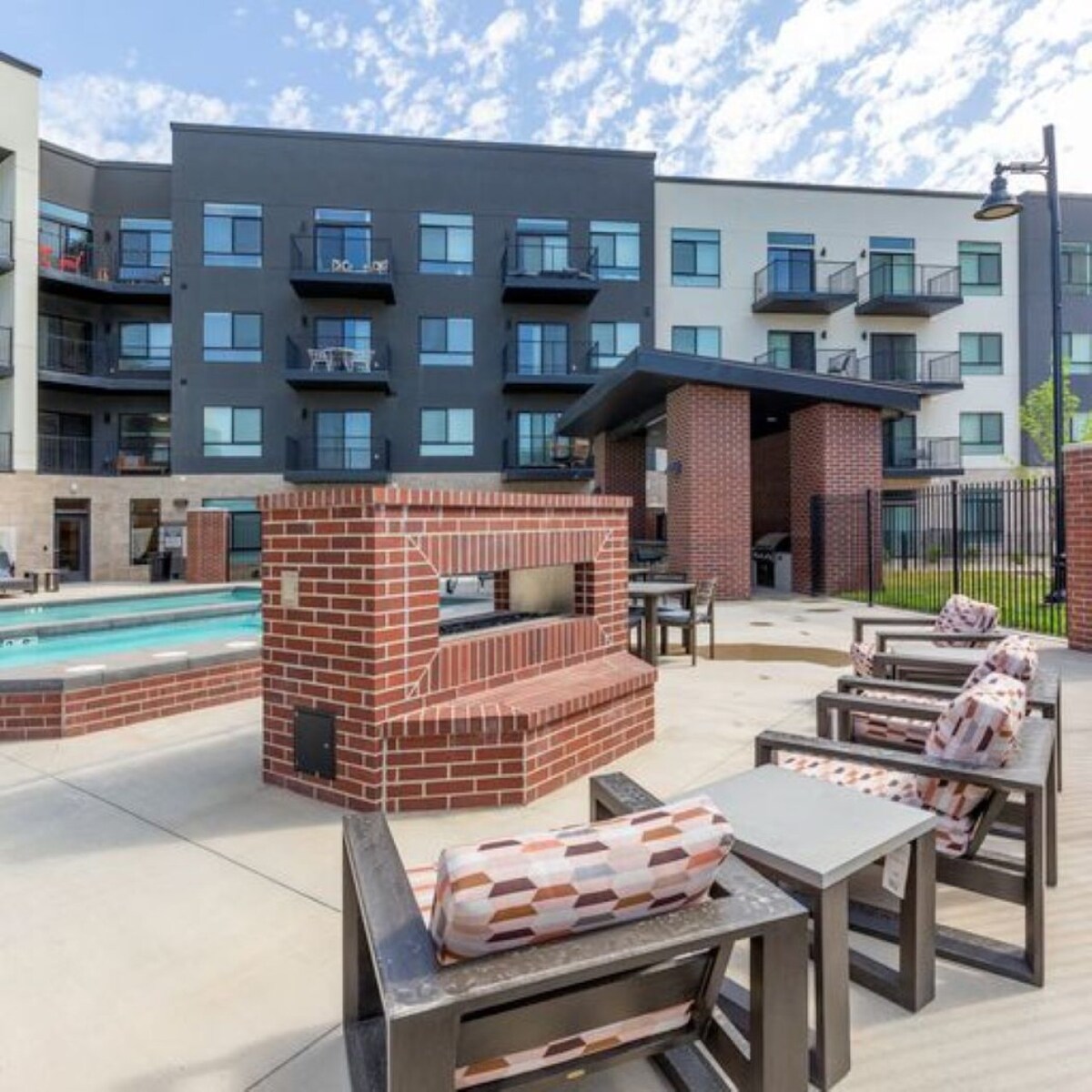 An outdoor space features a brick fireplace surrounded by modern seating, including comfort-focused chairs. The background displays a multi-story apartment building with balconies. Expansive concrete areas provide ample space for relaxation, complemented by a clear sky above.