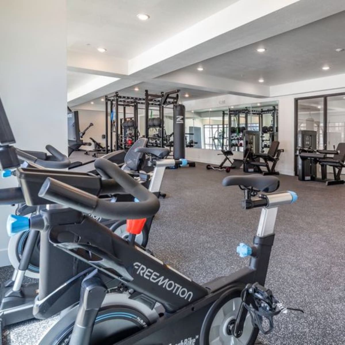 A modern gym space is filled with a variety of fitness equipment, including stationary bikes and weight machines. Bright lighting enhances the open layout, while mirrors line the walls, creating an expansive feel. Rubber flooring provides comfort and safety during workouts.