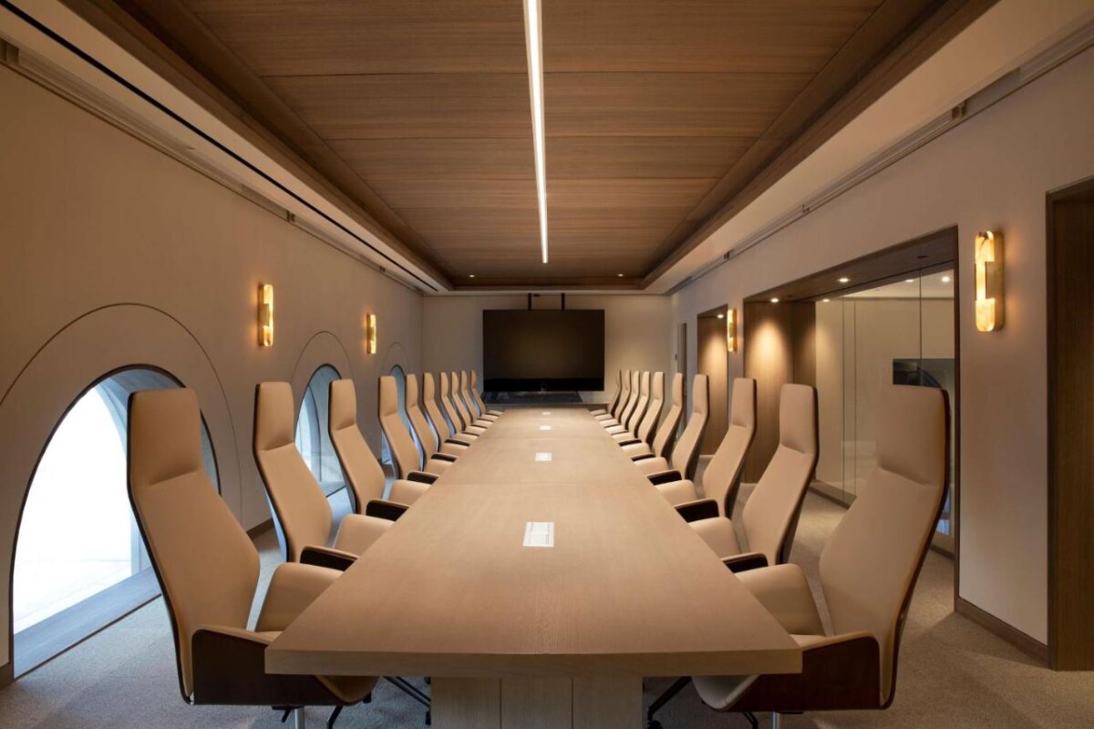 A modern conference room is equipped with a long wooden table, surrounded by comfortable tan chairs. Soft lighting is incorporated in the ceiling design, and a large flat-screen TV is mounted at the front. Arched windows add unique architectural details.