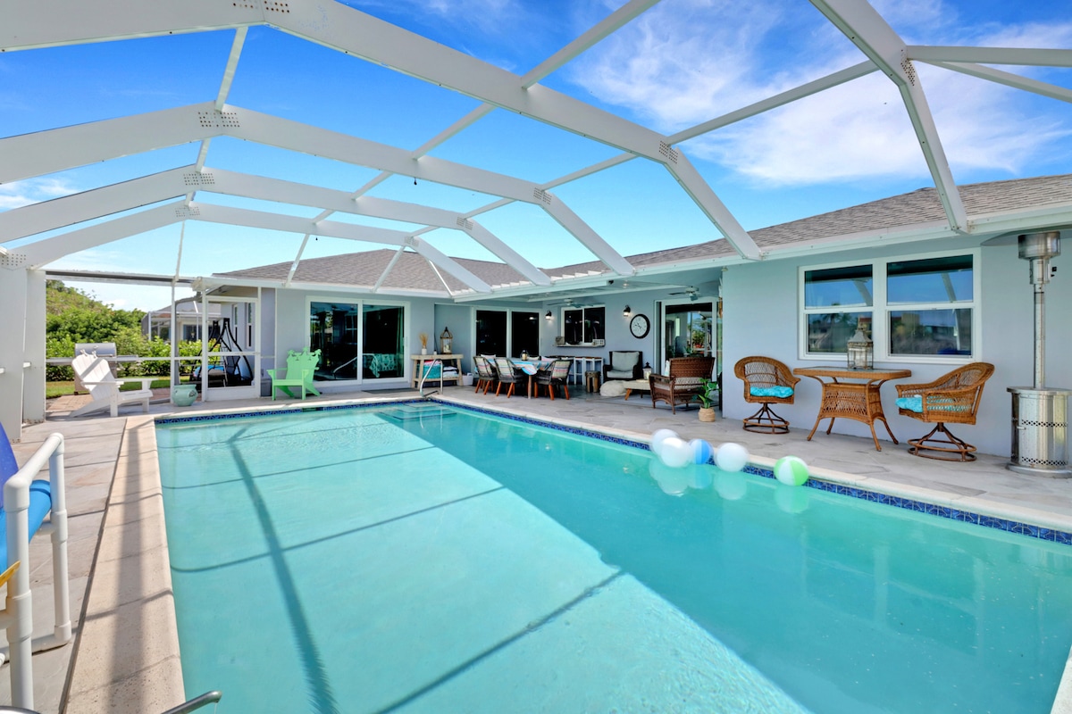 An inviting screened-in pool area features a shimmering pool surrounded by lounging chairs and outdoor seating. Sunshine streams through the mesh enclosure, highlighting the blue water and the lush greenery beyond. Colorful pool toys float gently on the surface.