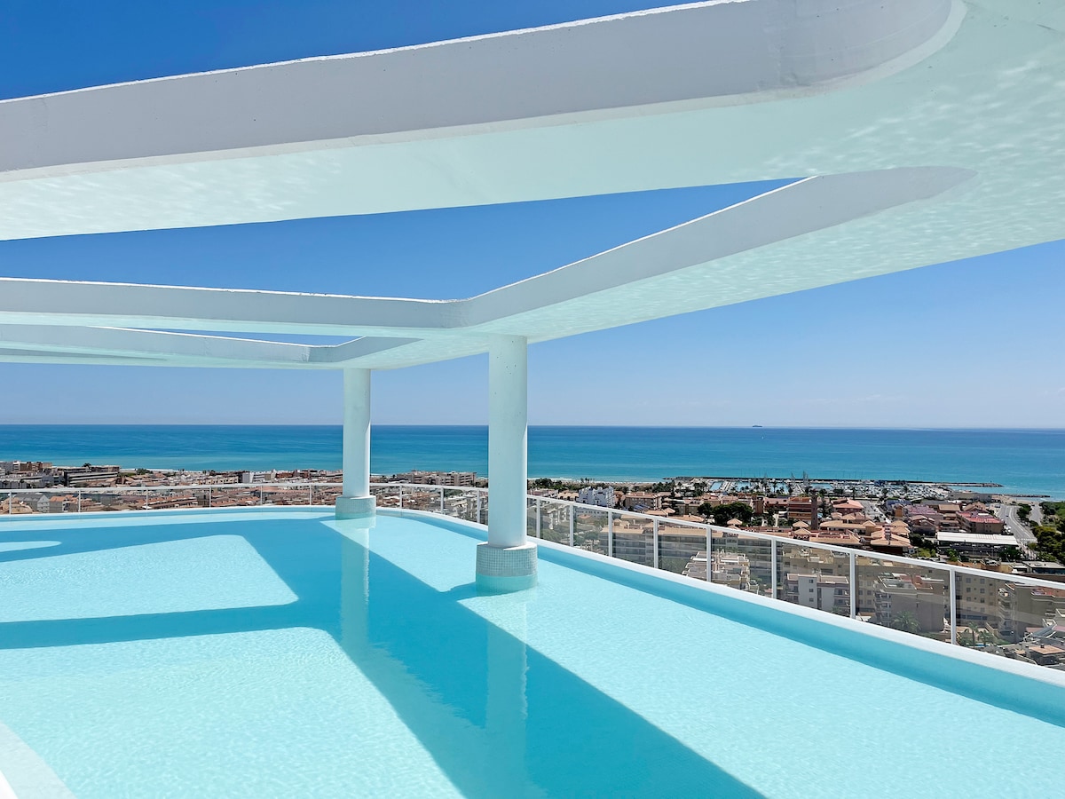 A serene infinity pool with clear turquoise water reflects the blue sky above. The pool area is surrounded by a modern white structure, offering expansive views of the coastline and the Mediterranean Sea in the distance.