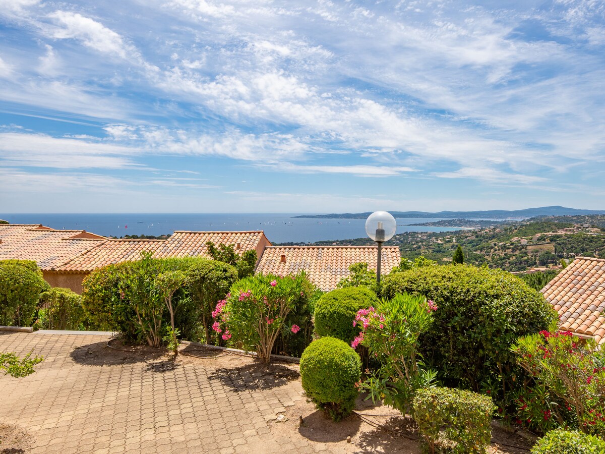 An expansive view encompasses the shimmering sea and lush valley, framed by well-maintained greenery and blooming flowers. The sky is bright with wispy clouds, and a pathway lined with hedges leads to the horizon, complemented by terracotta roofs in the foreground.