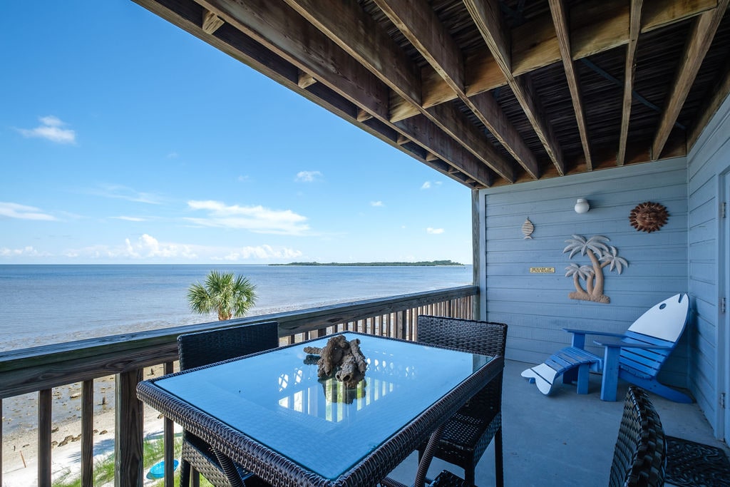 A balcony space is presented with a glass-top table surrounded by dark wicker chairs. Two colorful lounge chairs in blue are positioned nearby. The view of the open Gulf is visible, along with palm trees and a clear blue sky overhead.