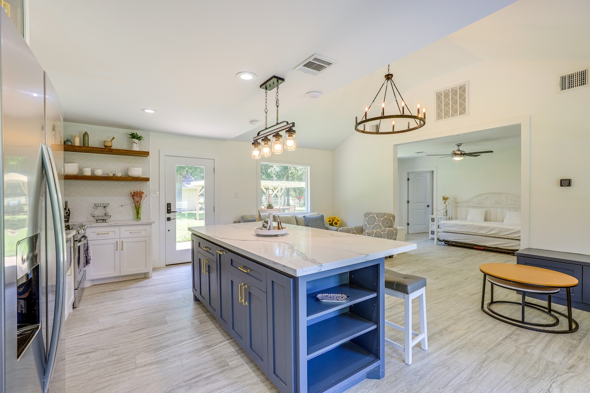 A modern kitchen features a central island with seating and a mix of dark blue cabinetry. Stainless steel appliances and light countertops are complemented by natural light from large windows. The adjoining living space, partially visible, includes a daybed and neutral-colored furnishings.