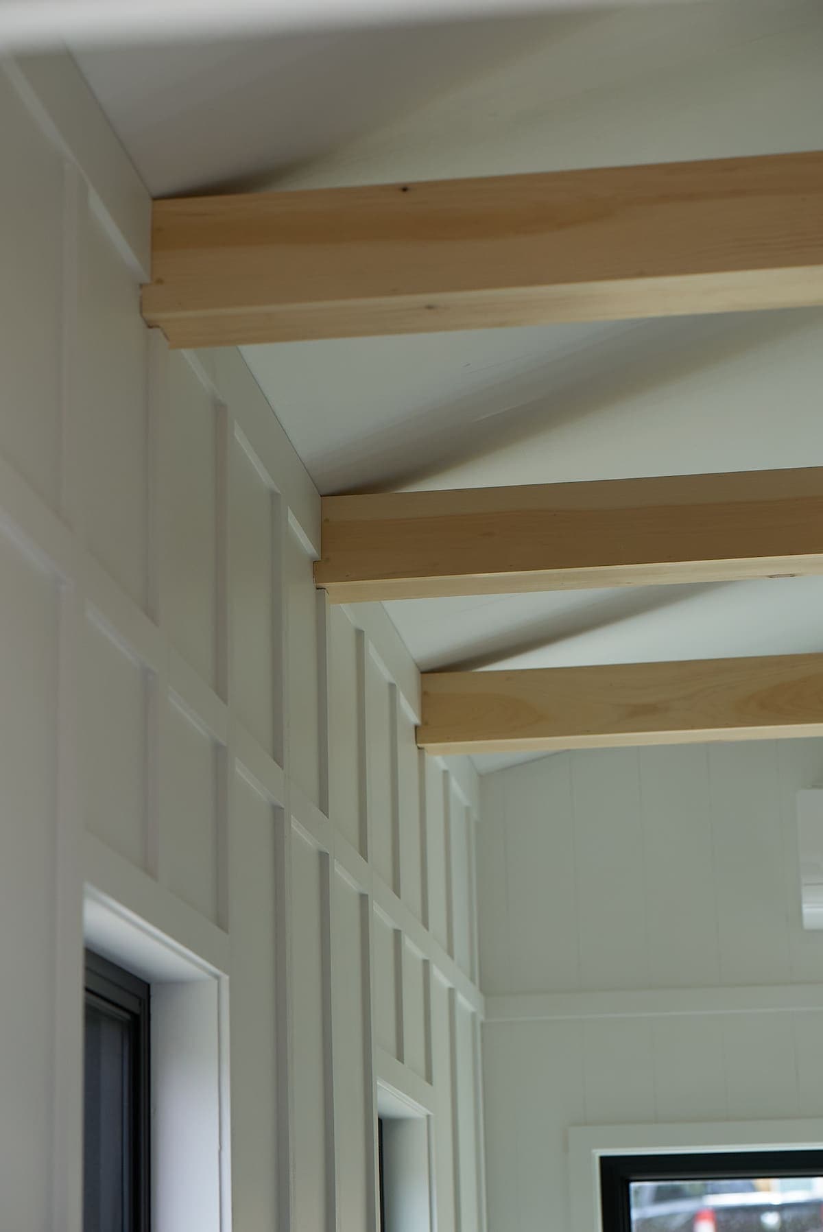 The image showcases a section of the interior where light wood beams stretch across the ceiling, enhancing the spaciousness of the area. The walls feature a clean, white paneling design, complemented by large black-framed windows that allow natural light to enter.