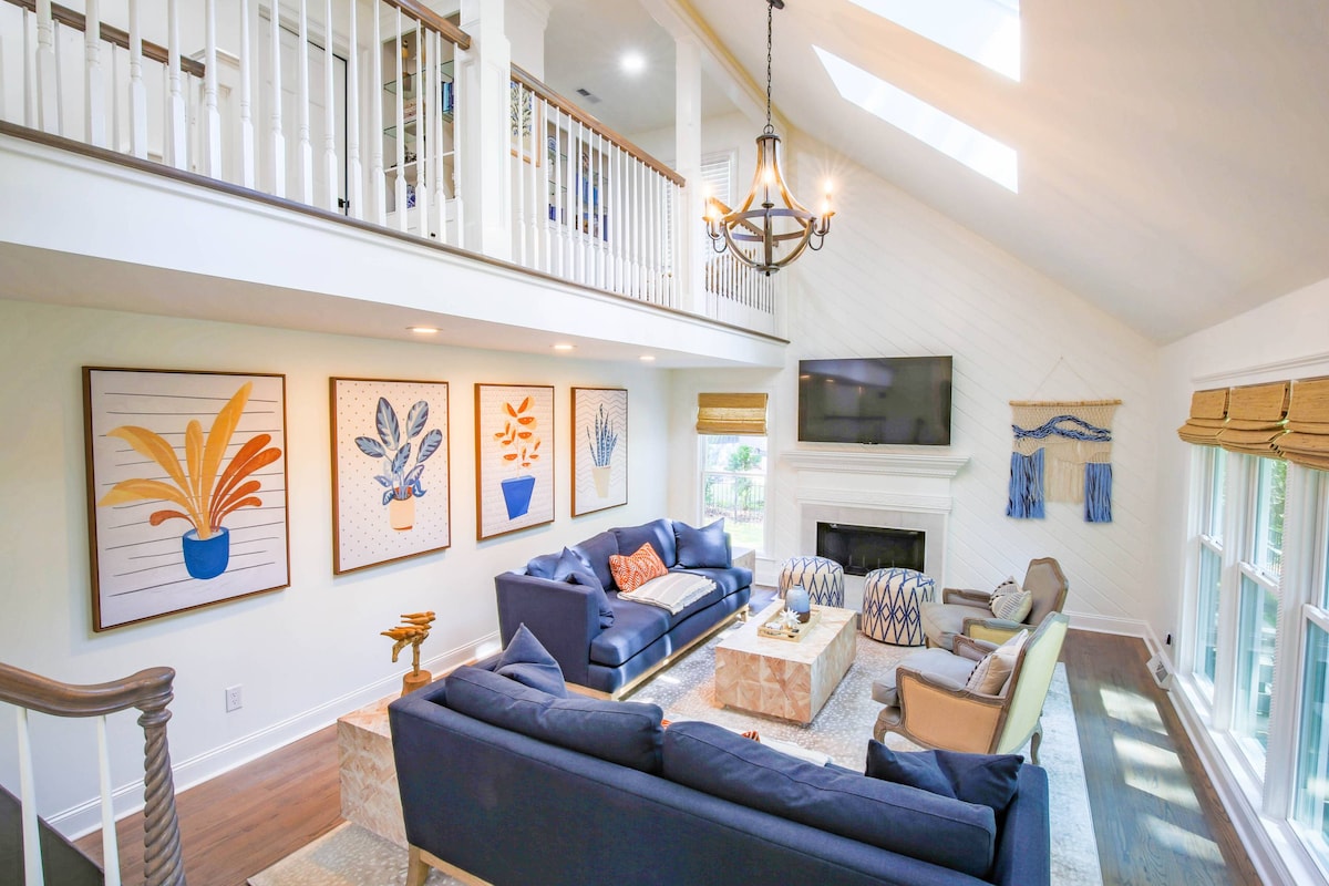 A spacious living area features a large sectional sofa in navy, complemented by accent chairs. Bright wall art depicting plants adorns the light-colored walls. Skylights and large windows enhance natural light, and a cozy fireplace adds warmth to the room's inviting atmosphere.