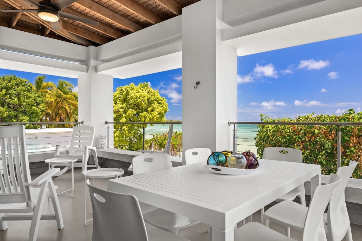 An outdoor balcony features a white dining table surrounded by white chairs, with vibrant views of the greenery and ocean beyond. The area is well-lit, showcasing the clear blue sky and inviting sea, enhancing the overall appeal of the space.