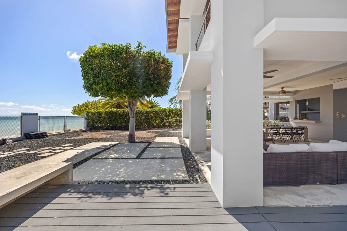An outdoor area showcases a spacious terrace with a neutral stone pathway leading to a well-maintained garden space. The ocean is visible in the background, framed by tropical greenery. Ample seating is provided for relaxation and dining.