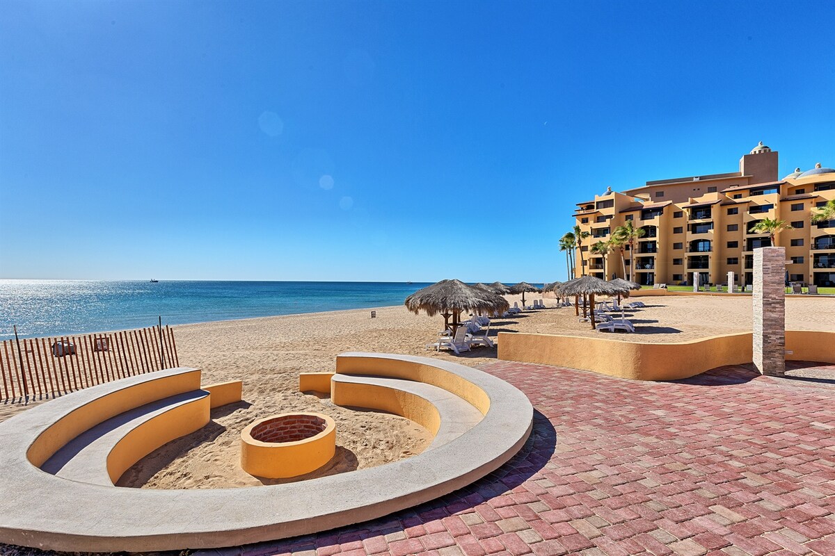 A circular fire pit is set in a sandy area facing the beach, surrounded by a paved pathway. Palapas with lounge chairs are visible on the shore, and the ocean stretches out in the background under a clear blue sky.