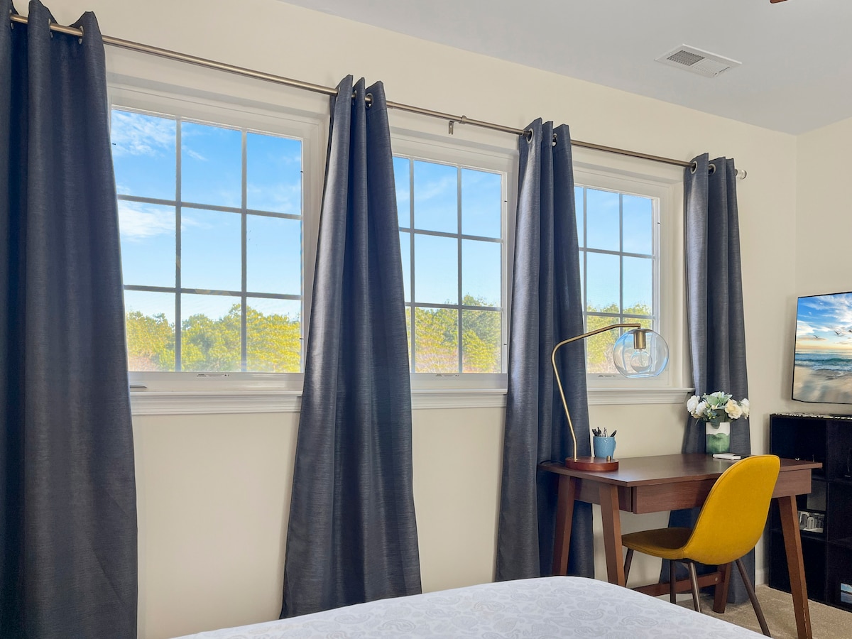A private bedroom features large windows adorned with dark curtains, allowing natural light to fill the space. A stylish desk with a yellow chair is positioned nearby, complemented by a lamp and decorative plants. A flat-screen TV is visible, providing entertainment options.
