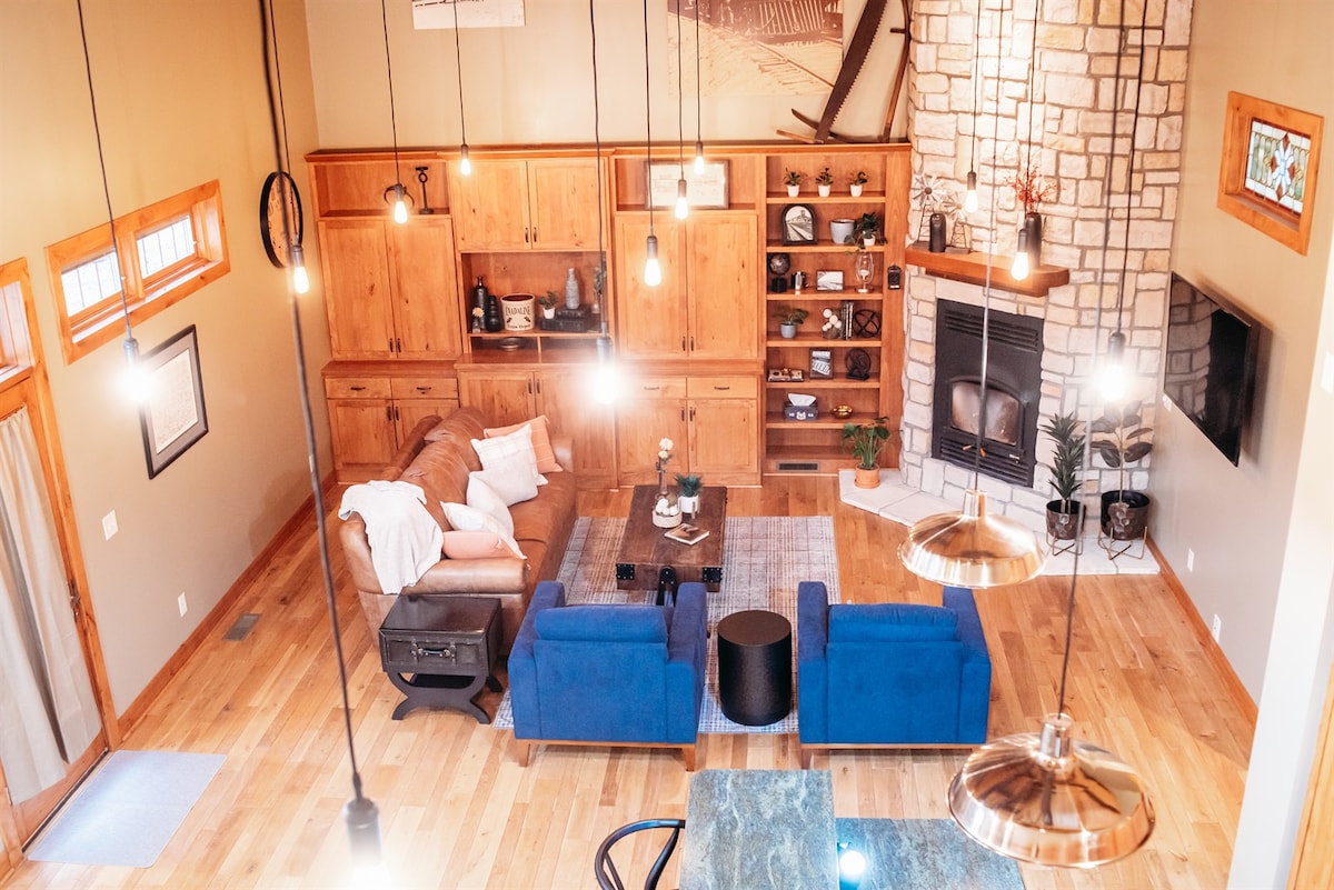 An open living space features a warm wood cabinetry and a stone fireplace as focal points. Soft lighting enhances the seating area, which includes a sofa and two blue chairs. A coffee table is centered on a textured rug, with decorative items elegantly arranged on shelves.