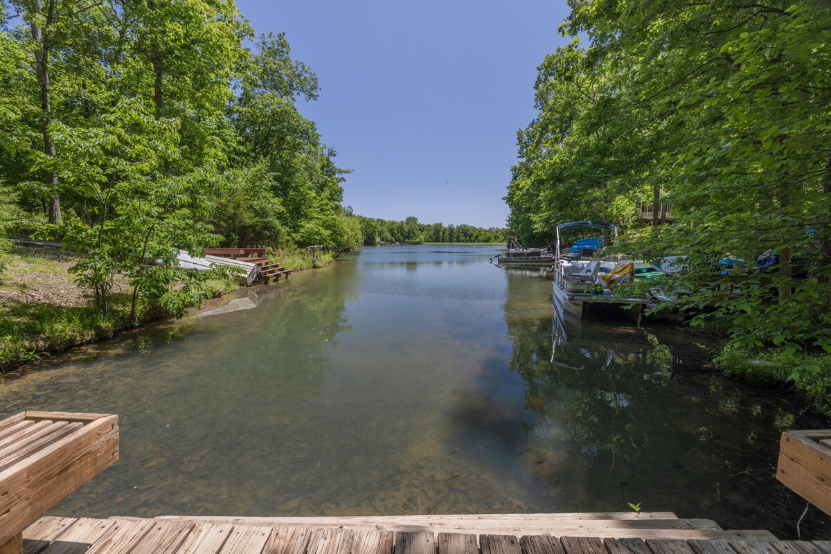 A tranquil view of the lake is captured from the wooden dock, surrounded by lush greenery. The clear water reflects the blue sky, while nearby boats are visible along the shoreline, enhancing the peaceful lakeside atmosphere.