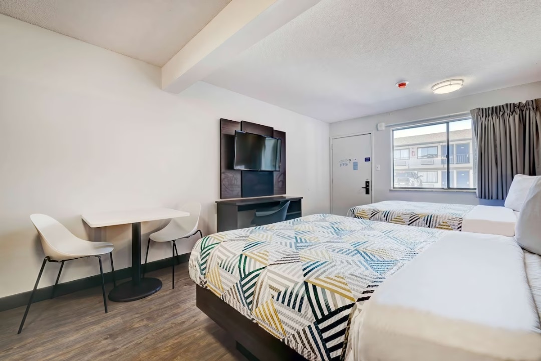 The room features two queen beds, each adorned with patterned bedding. A small dining table with two chairs is positioned near the window, allowing natural light to fill the space. A wall-mounted television is installed above a storage unit across from the beds.