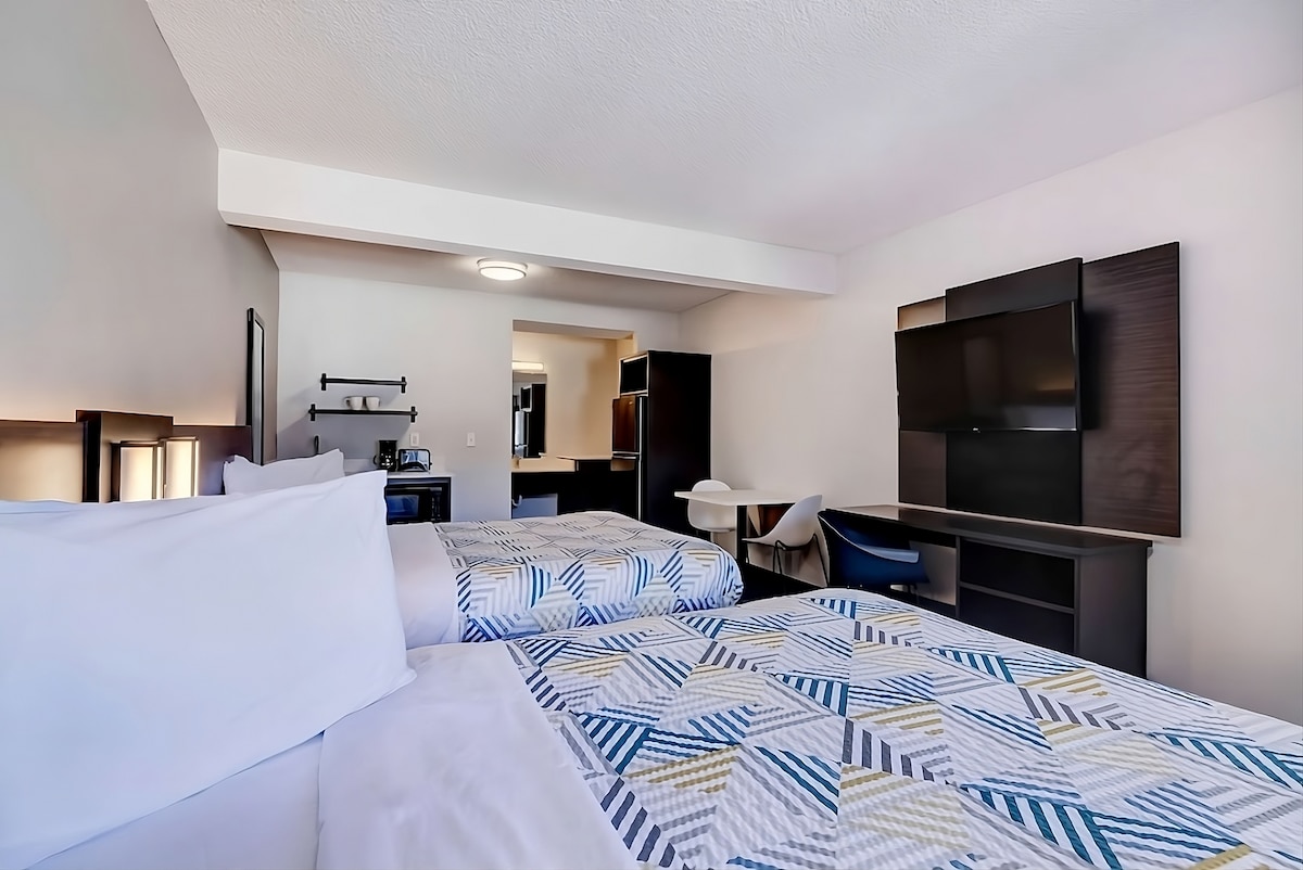 A well-organized hotel room features two Queen beds with patterned covers. A work desk and chair are positioned near a flat-screen TV. The kitchenette, visible in the background, includes essential appliances. Ample lighting enhances the functional design of the space.
