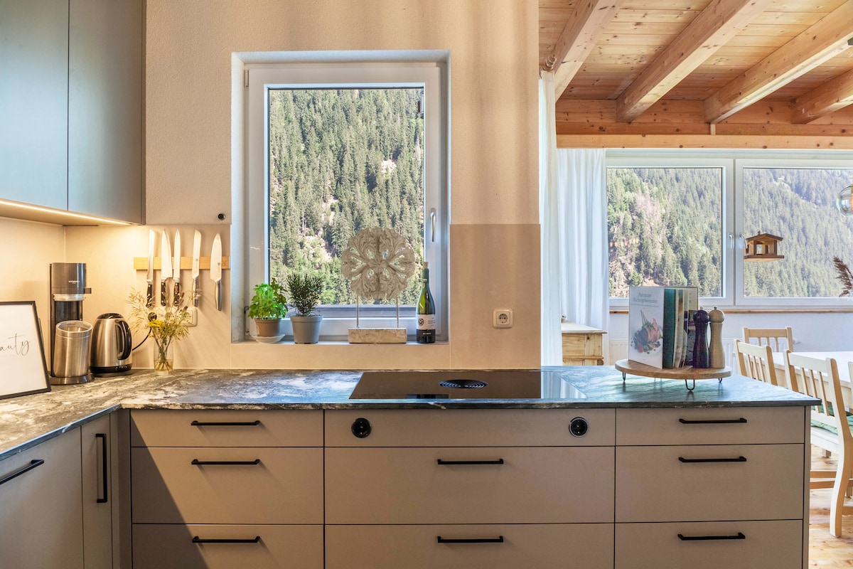 A modern kitchen with a granite countertop is equipped with appliances, including a coffee maker and kettle. A window offers a view of the surrounding mountains, and natural light brightens the space. Potted plants and decorative items add a touch of greenery and style.