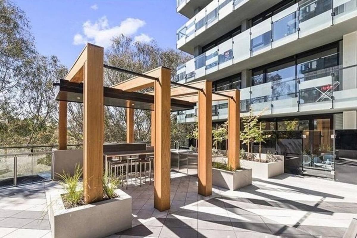 An outdoor dining area is presented with a wooden pergola, featuring a built-in table and benches. Planters with greenery surround the space, which opens up to the building's modern architecture and offers a tranquil atmosphere for guests to enjoy meals outdoors.