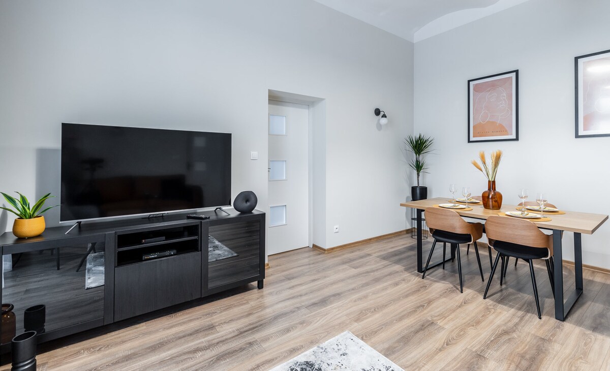 A modern living area features a large television mounted on a dark entertainment unit. A wooden dining table is set for four, with minimalist table decor. Light wood flooring adds warmth, complemented by a potted plant and framed artwork on the walls.