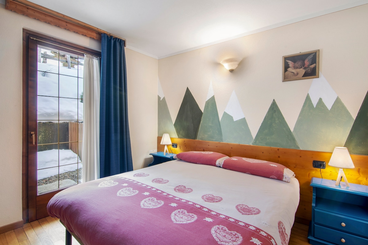 A comfortable bedroom features a large bed adorned with a patterned blanket. Mountain-themed murals add a touch of nature, while bedside tables with lamps provide gentle lighting. A glass door opens to a snowy outdoor area, enhancing the serene atmosphere.