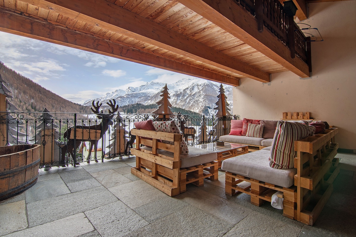 A spacious balcony features comfortable seating arrangements made from wooden pallets, adorned with cushions in warm tones. Scenic mountain views and decorative elements like wooden trees create a relaxed atmosphere, while the stone floor adds a touch of rustic charm.