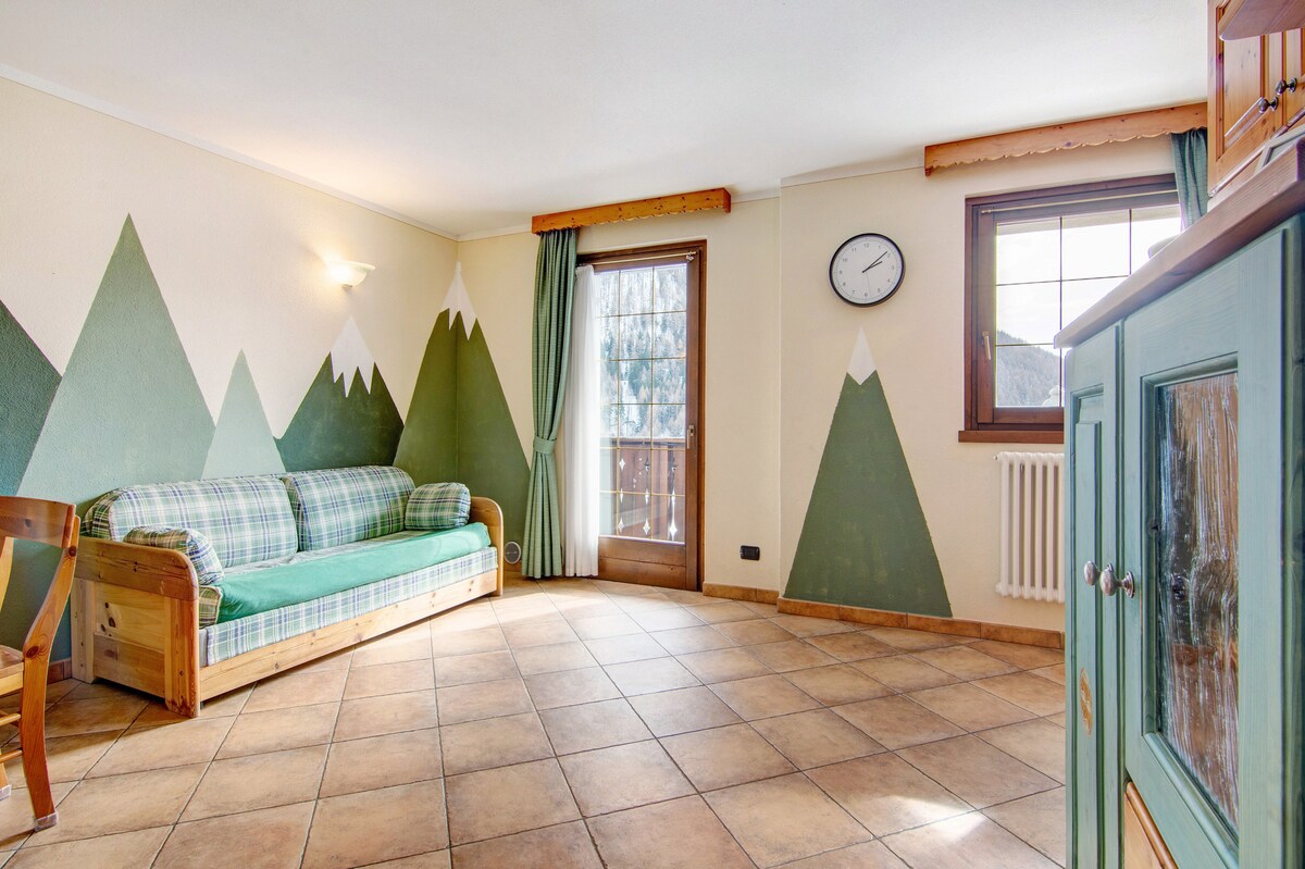 A spacious living room features a green sofa bed, adorned with a plaid design. The walls are decorated with mountain motifs, creating a natural ambiance. Large windows invite ample light, while a wall clock and radiator complete the room's functionality.