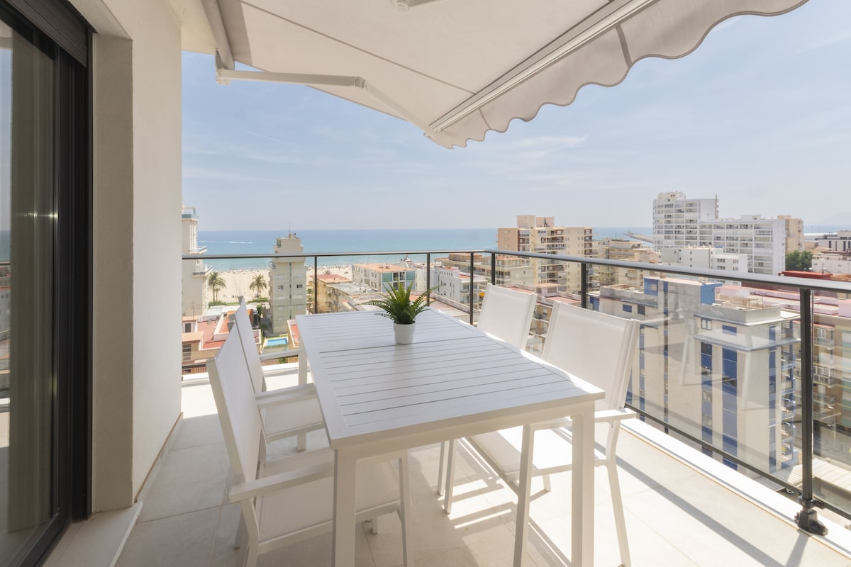 A sunlit roof terrace features a white outdoor dining table surrounded by several chairs. An umbrella provides shade, while the sea views extend into the distance. The contemporary design includes a railing overlooking city buildings and the coastline.
