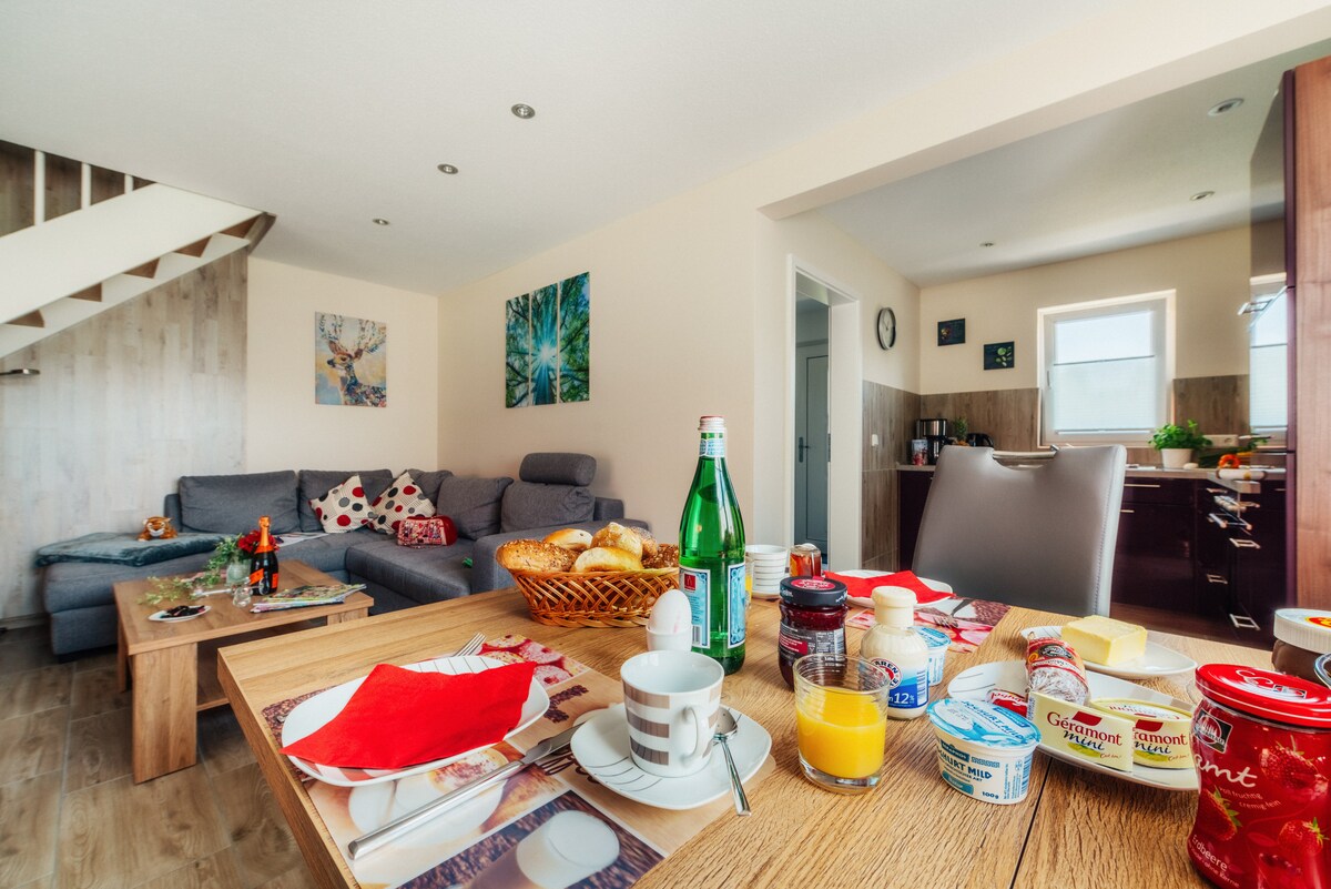 A living and dining area features a comfortable double sofa bed, a wooden dining table set with breakfast items, and decorative wall art. The modern kitchen is visible in the background, complete with a window that brings in natural light.