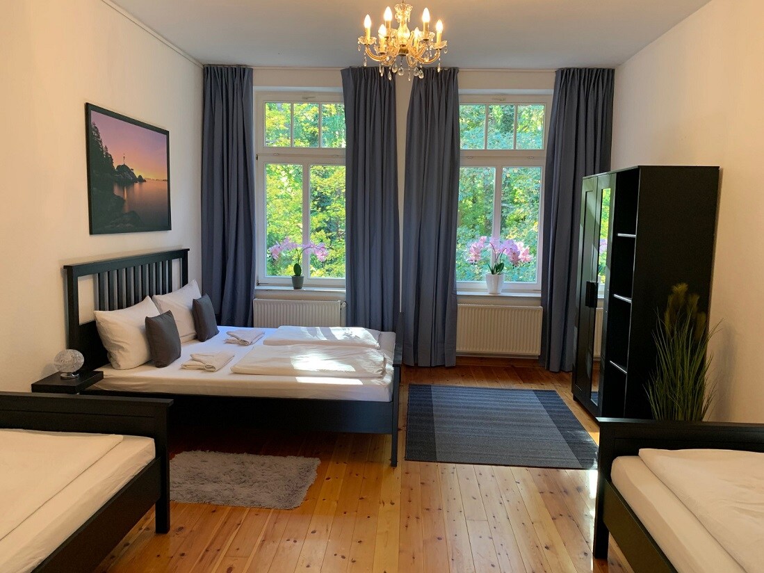 A spacious sleeping area is highlighted by large windows adorned with flowing curtains, allowing natural light to fill the room. Two beds are positioned alongside a chest of drawers, and a decorative rug complements the wood flooring.