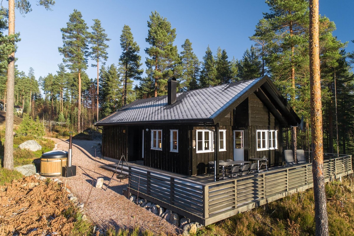 A charming chalet is situated among tall trees, featuring a natural wood exterior. A spacious deck with outdoor seating offers a relaxed setting, while a hot tub rests nearby. The pathway leading up to the chalet is lined with gravel, enhancing the tranquil environment.