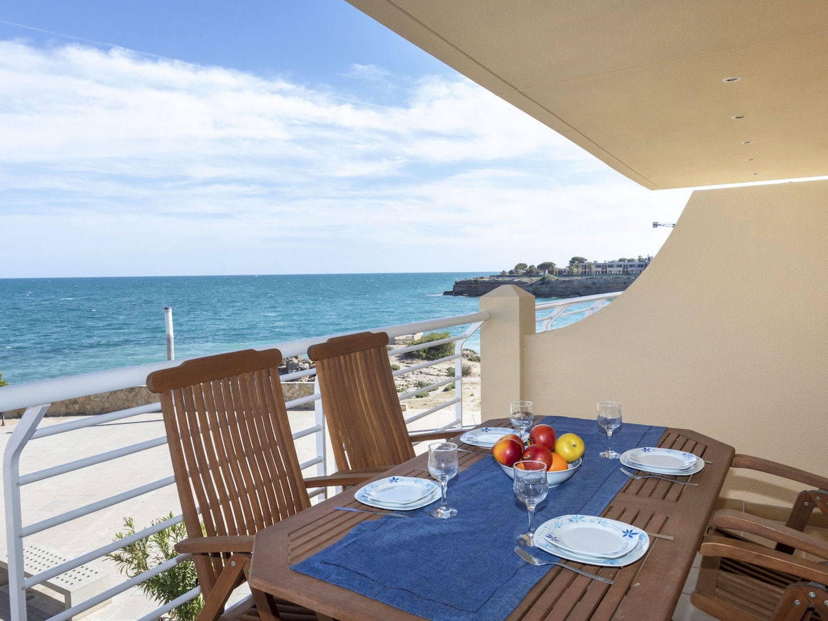A terrace dining area is shown with wooden chairs and a table set for four, featuring a blue tablecloth. Fresh fruit is displayed on the table alongside glassware, with a view of the sea and coastal landscape visible in the background.