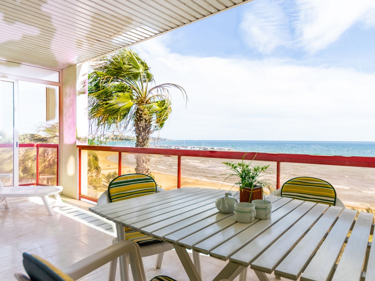 An outdoor terrace is furnished with a white table and comfortable chairs, offering a view of the ocean and the beach. Green palm trees sway gently in the breeze, and the sky is filled with soft clouds, creating a serene atmosphere.