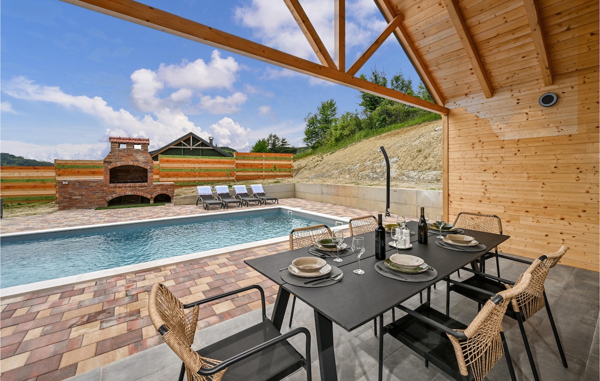An outdoor terrace features a modern dining area with a dark table and stylish chairs, set against a backdrop of a private swimming pool. Sun loungers are arranged beside the pool, and a stone barbecue is visible in the distance, framed by a wooden structure.