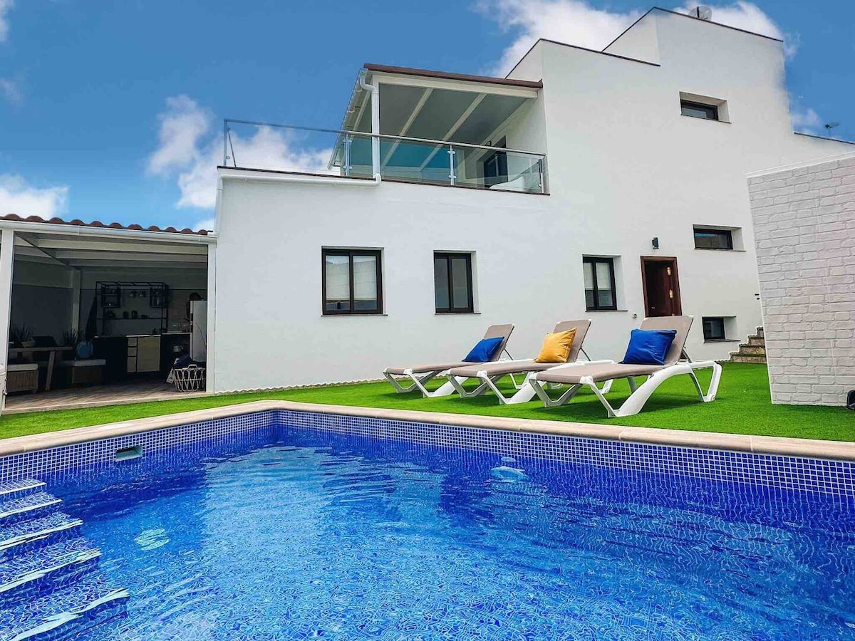 A modern villa features a sparkling blue pool accompanied by white lounge chairs adorned with colorful cushions. The two-story structure exhibits clean lines and a minimalist design, with large windows offering ample natural light. A covered outdoor seating area is visible in the background.