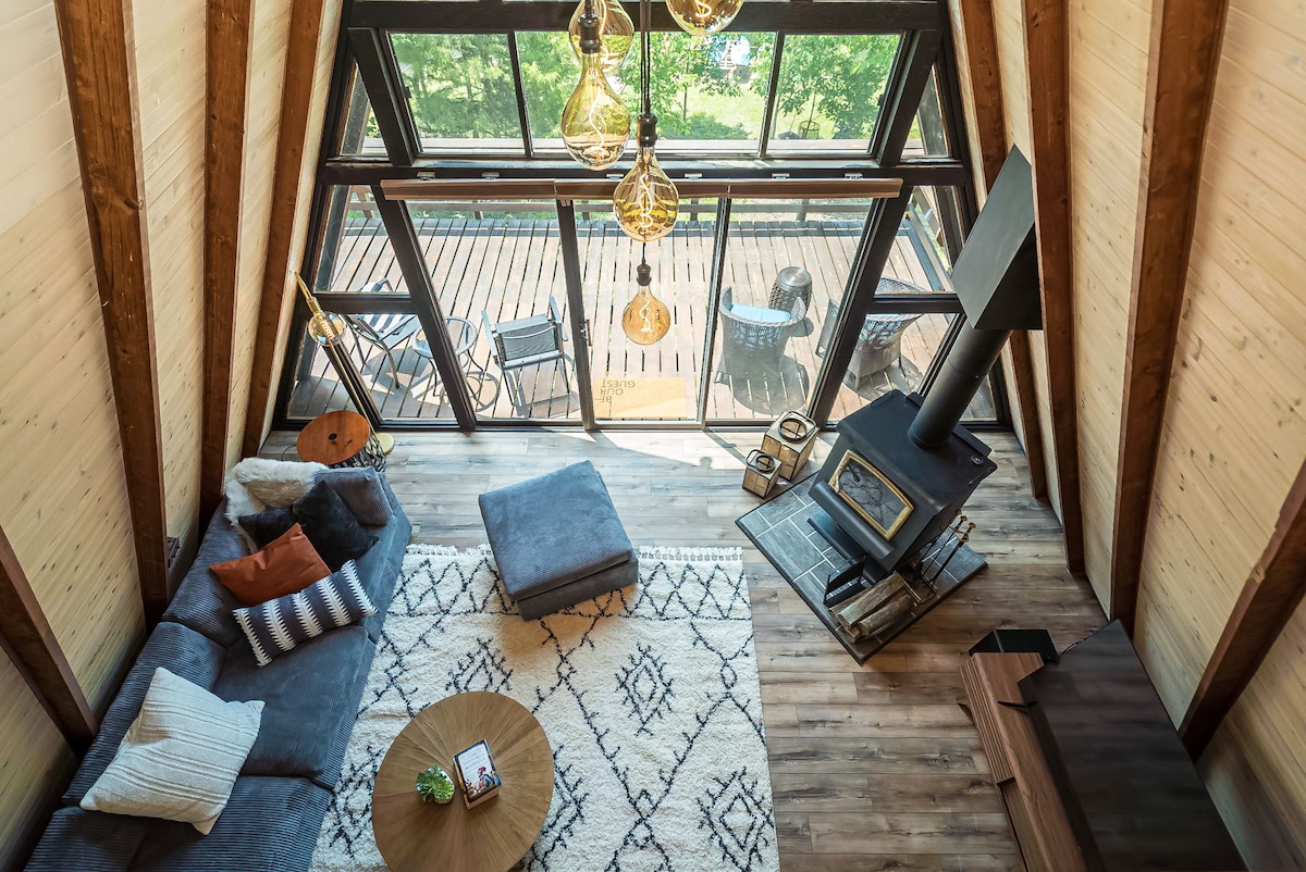 An airy, open living space is highlighted by large windows allowing natural light to stream in. A wood-burning stove is positioned beside a cozy seating area featuring plush cushions and a patterned rug. A round coffee table sits in the center, creating a welcoming atmosphere.