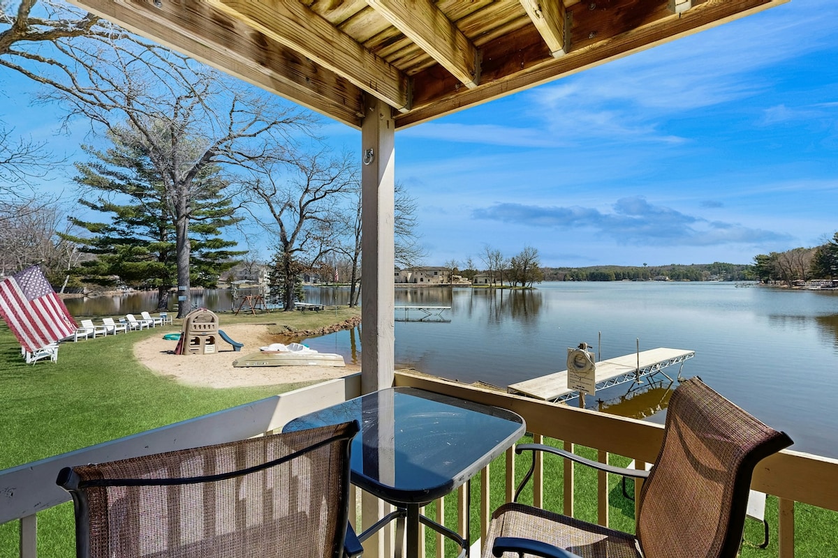 A private deck overlooks the calm waters of Lake Delton, featuring two chairs and a small table. In the distance, sandy beach and trees provide contrast against the clear blue sky, while a wooden dock extends into the lake, inviting tranquil moments in nature.