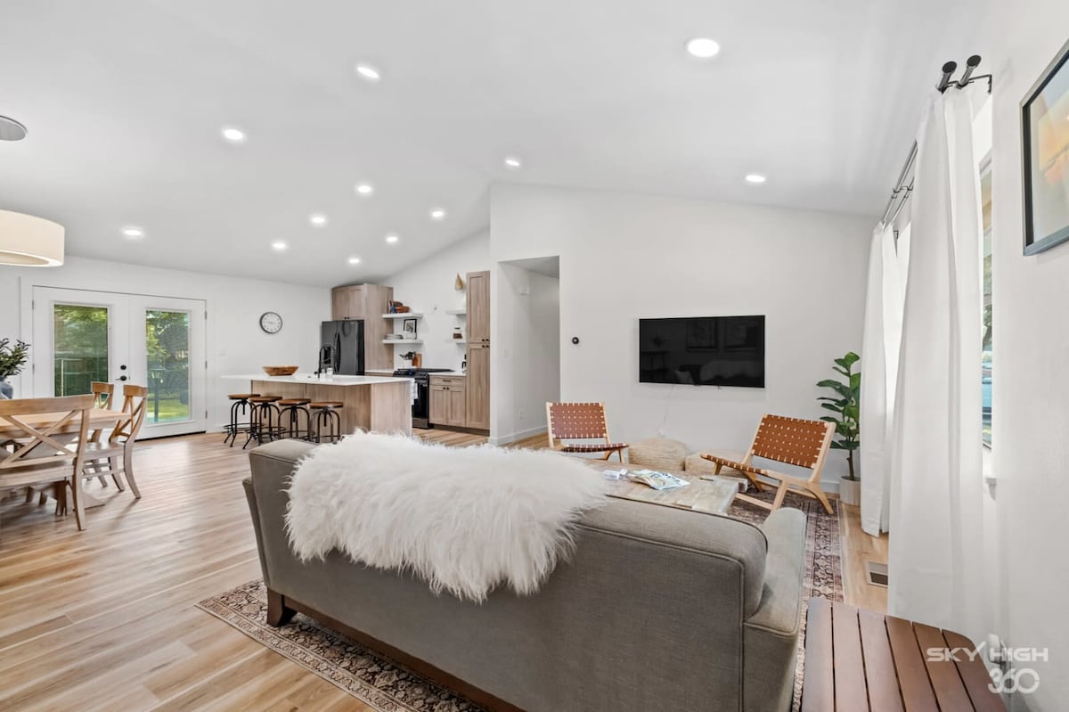 A bright and airy living space features modern furnishings with a cozy couch accented by a white throw. A wooden dining table accommodates several chairs, while a well-equipped kitchen is visible in the background. Natural light fills the room from sliding glass doors.