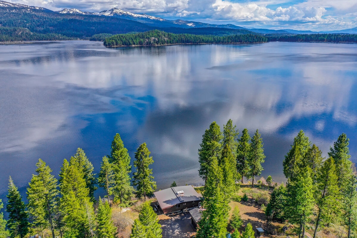 An aerial view captures the Laur House surrounded by lush trees, with the expansive Little Payette Lake reflecting the sky. The serene water contrasts beautifully with the greenery, while distant mountains loom beneath a partly cloudy sky.