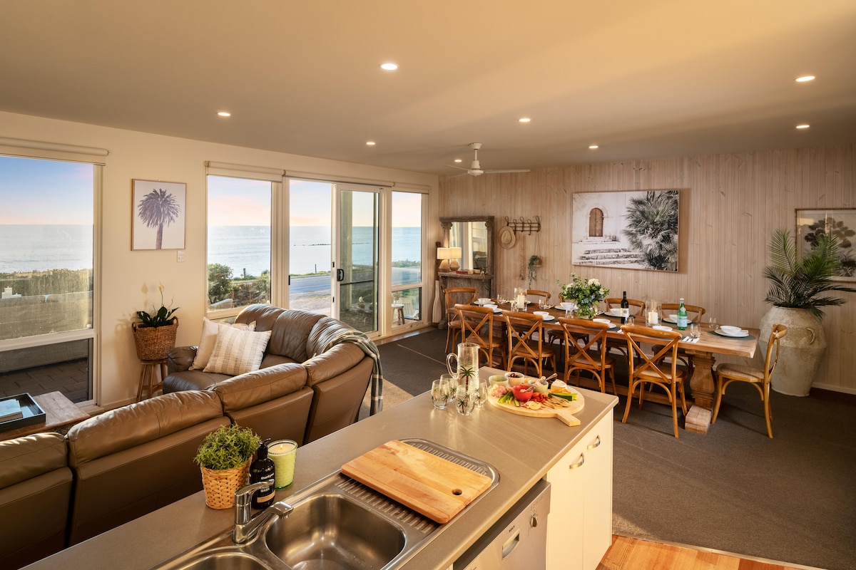 The open-plan living area showcases a spacious kitchen with a sink and preparation area, transitioning into a dining space equipped with wooden tables and chairs. Large glass doors reveal ocean views, while natural light enhances the contemporary decor of the lounge, featuring comfortable seating.