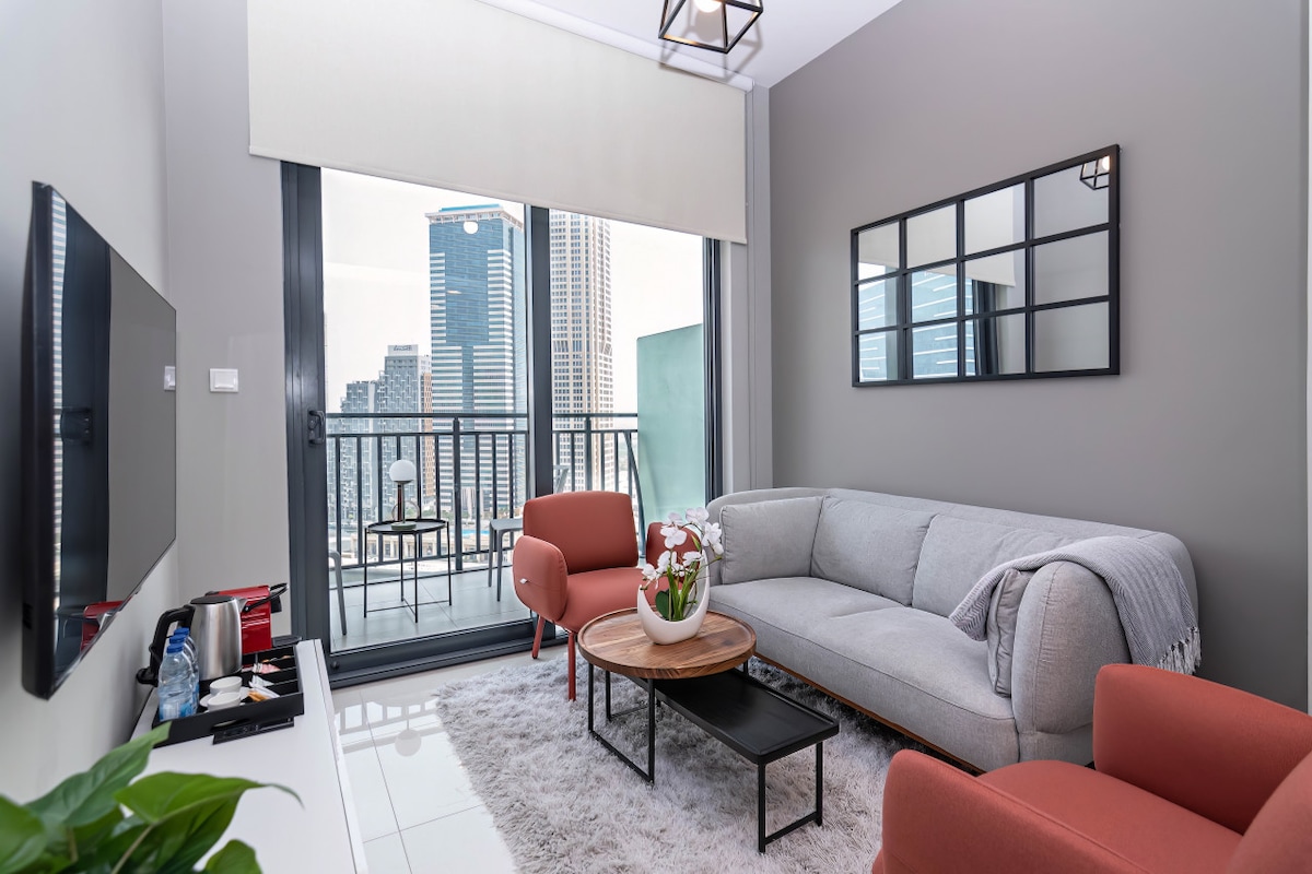 A contemporary living area features a comfortable gray sofa paired with two orange chairs. A round coffee table sits in front, with a sleek flat-screen TV mounted on the wall. Large windows provide city views and natural light, while a small balcony is visible.