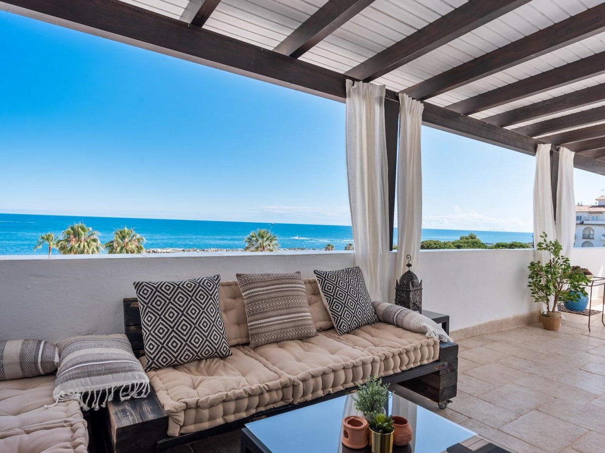 A spacious terrace is featured, showcasing a comfortable seating area with plush cushions and neutral-toned decor. The sea view is unobstructed, complemented by palm trees in the background. Sheer curtains gently frame the space, while a glass table holds decorative items.