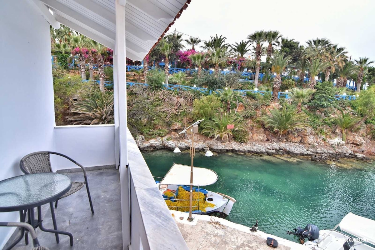 A private balcony with a small round table and two chairs overlooks a serene waterway. Lush greenery and palm trees line the opposite shore, offering a relaxing natural backdrop. A small boat is anchored nearby, complementing the tranquil waters.