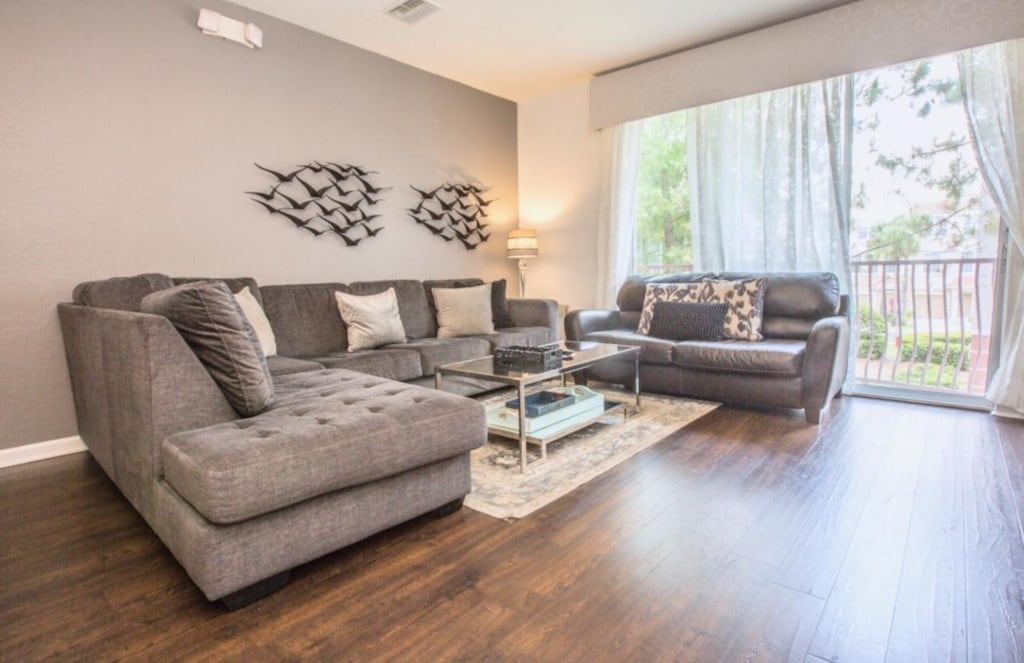 A spacious living area features hardwood flooring, complemented by a large gray sectional sofa and a black leather sofa. A glass coffee table sits at the center, with decorative accents noted on the walls. Natural light filters in through sheer curtains, enhancing the welcoming ambiance.
