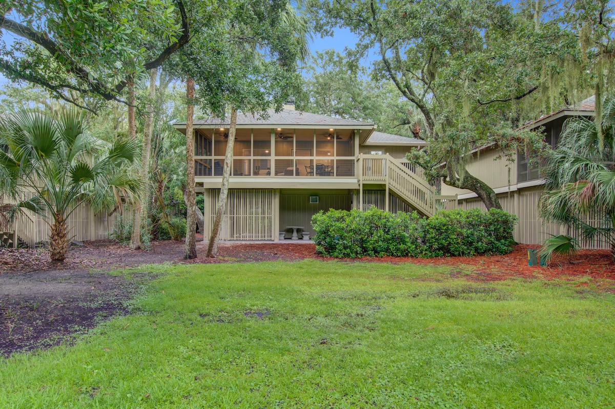 The exterior view showcases a home surrounded by lush greenery and mature trees. The screened-in porch extends from the upper level, providing a shaded area. A spacious lawn is visible in the foreground, bordered by landscaping that includes palm plants.