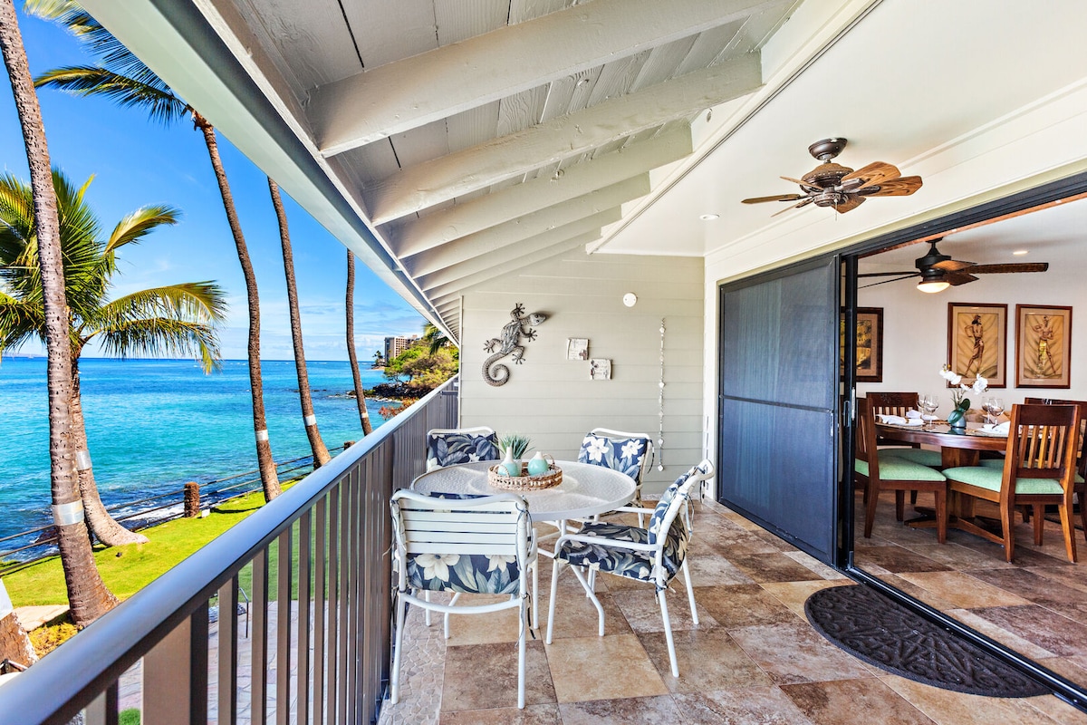 An expansive lanai features a round dining table surrounded by four chairs, overlooking vibrant ocean waters. Tropical landscaping and palm trees frame the view, while sliding glass doors offer access to the living area. Natural light brightens the space, enhancing the coastal ambiance.