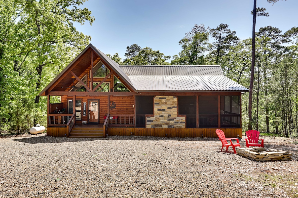 A charming cabin is nestled among the trees, featuring a peaked roof and wooden exterior. The cabin is complemented by a gravel area and a fire pit surrounded by seating. A screened porch invites relaxation, while vibrant red chairs provide a pop of color.
